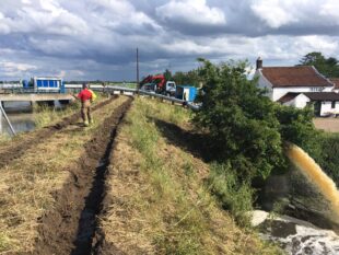 Environment Agency staff standing on a raised grassy road with a large pump gushing water at the base of the bank below them.