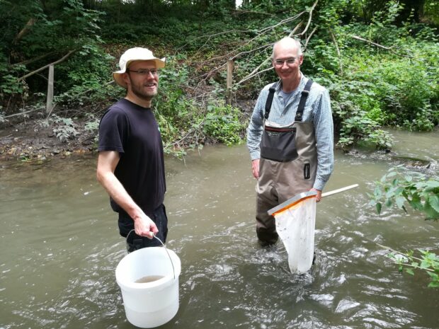 photo of two people stood in a river with a bucket and a fishnet to do some monitoring