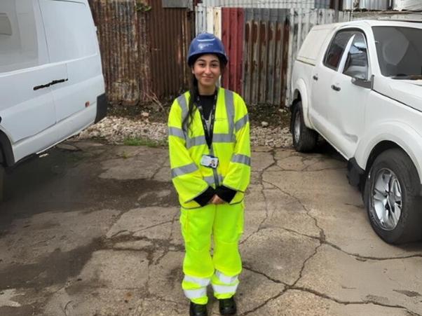 An Environment Agency officer dressed in yellow high-vis and blue helmets stands in front of a large, corrugated metal fence