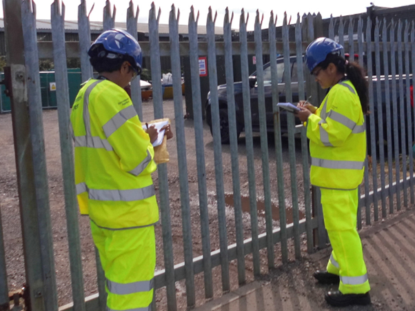 Two Environment Agency interns dressed in yellow high vis trousers and jackets, and blue helmets make notes whilst standing in front a large metal fence in front of a regulated site. 