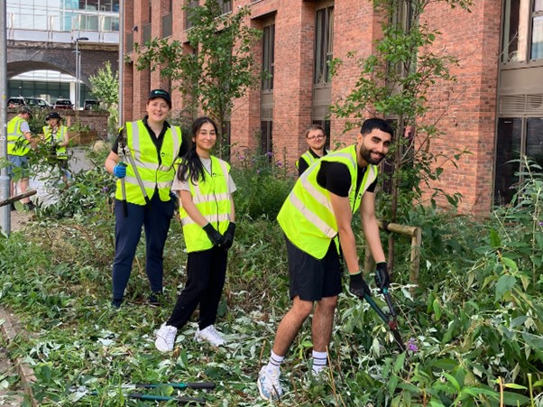 Six people from the Environment Agency dressed in yellow high-vis jackets are surrounded by pruned foliage, in front of a large red-bricked building. 