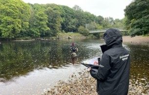 The image shows the River Wharfe at Cromwheel, with an Environment Agency officer in the water taking a water sample, and another standing on the river bank taking notes. 