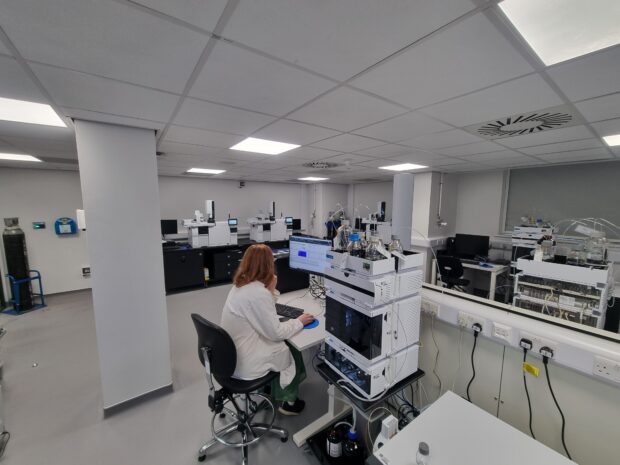 A scientist sits at a liquid chromatography station, working on the computer. Behind them, the modern laboratory floor is visible, featuring new analytical equipment and workstations in a bright, open plan setting