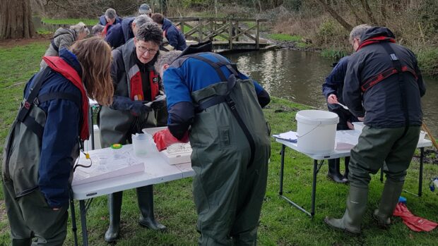 A group of people standing around tables at the riverbank carrying out Riverfly training.