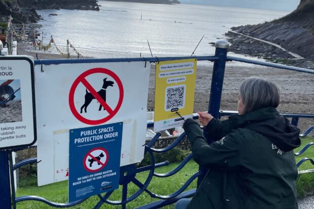A female staff member of the Environment Agency is seen attaching a QR code printed on thick plastic alongside other signs on metal railings in front of Combe Martin beachfront.