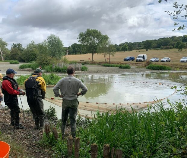 Three people in overalls look out towards a shallow body of water with a large net.
