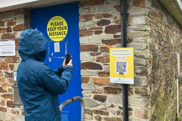 A person wearing a raincoat is seen pointing their smart phone at QR code signate attached to the wall of a brick building in a car park by the Par Sands beach.
