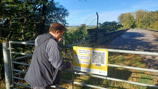 A man attaching a QR sign to a wide metal gate across the road by the River Nidd.