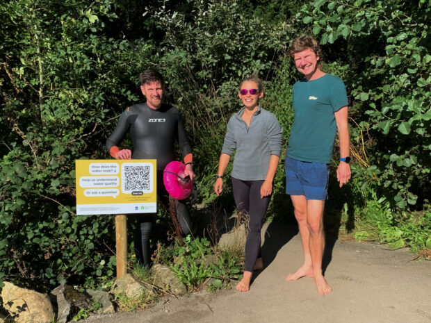 Three swimmers, two men and one woman, wearing a mixture of swimsuits, shorts and T-shirts, stood on the bank of Lank Windermere next to a QR sign post.