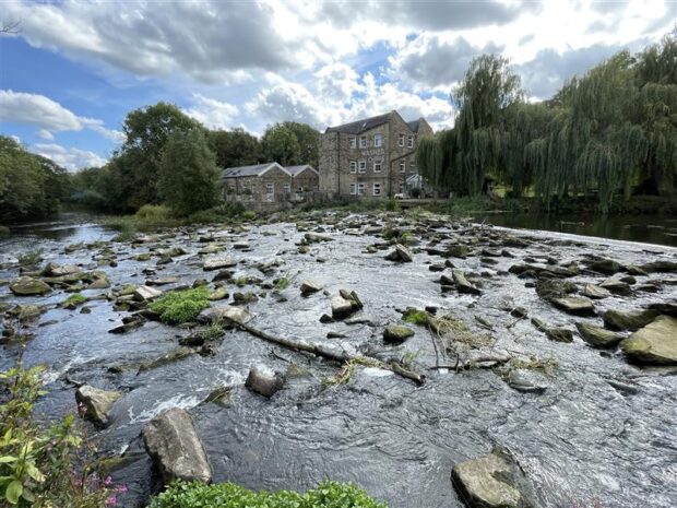 A photo of the River Aire in Yorkshire with Hirst Mill in the background. There are rocks in the river, known as rock ramps, which help fish move up the river.