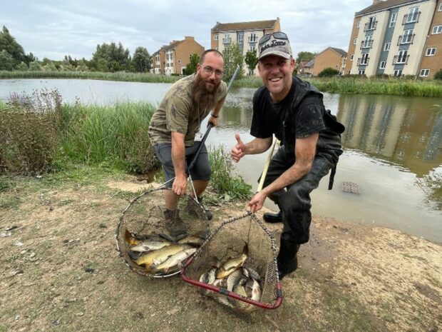 Two male Environment Agency officers looking at camera on a riverbank, each with a large handled net filled with fish.