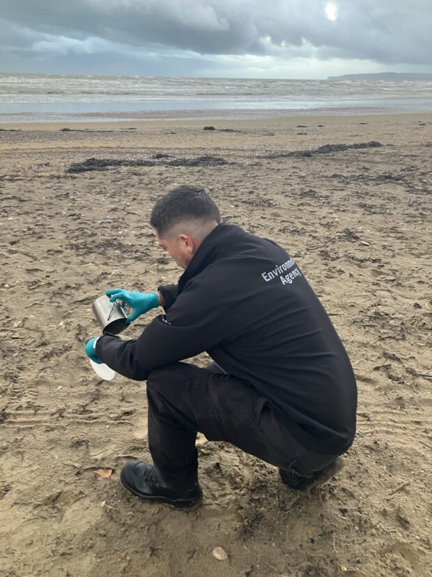 An Environment Agency officer kneels in the sand to collect samples