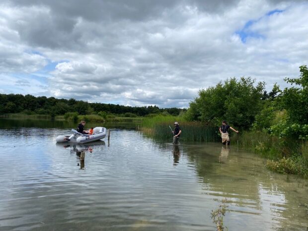 Three Environment Agency staff knee deep in a lake, one in a dinghy, collecting samples.