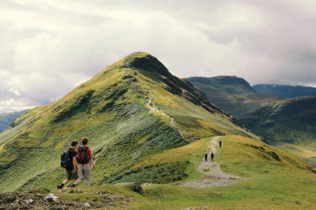 People hiking along a winding path up a grassy green hill.