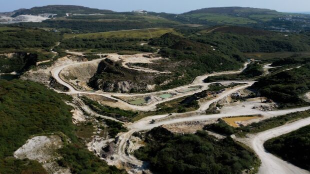 Aerial view of a china clay quarry showing dirt roads, pale rock faces, and pools of water. Surrounding the quarry, green vegetation and rural farmland stretch into the background.