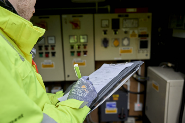A man in high vis marks down notes on his clipboard while looking at technical equipment.