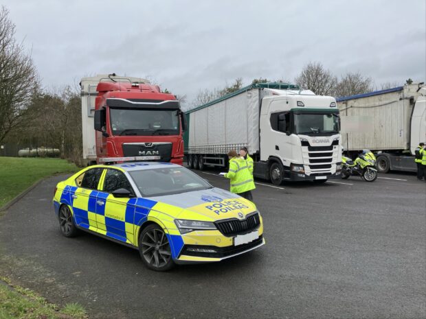 Image shows a parking area with a police car and bike and three lorries parked. Three officers in high visibility clothing are stood outside the vehicles.