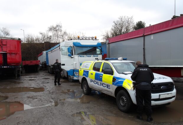 The image shows two police officers and a police truck. There is a lorry blocked by the police truck and other lorry trailers can be seen either side.