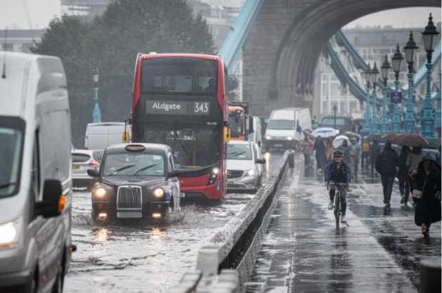 A London taxi, bus and cyclist travelling through flood water on Tower Bridge.