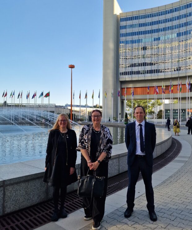 • Jo Nettleton standing with two colleagues beside fountains and international flags outside the International Atomic Energy Agency headquarters in Vienna.