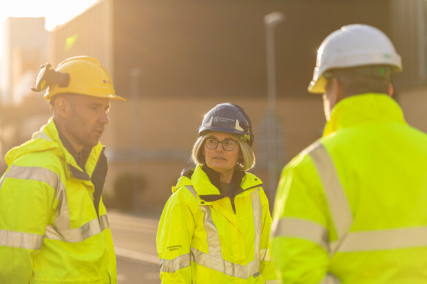 A female regulator wearing high-visibility clothing and a hard hat while speaking with two colleagues at a nuclear site.