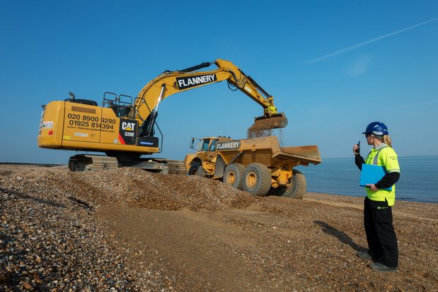 A digger dropping shingle into a truck to be spread on a beach. An Environment Agency officer talks on a walkie talkie next to it.